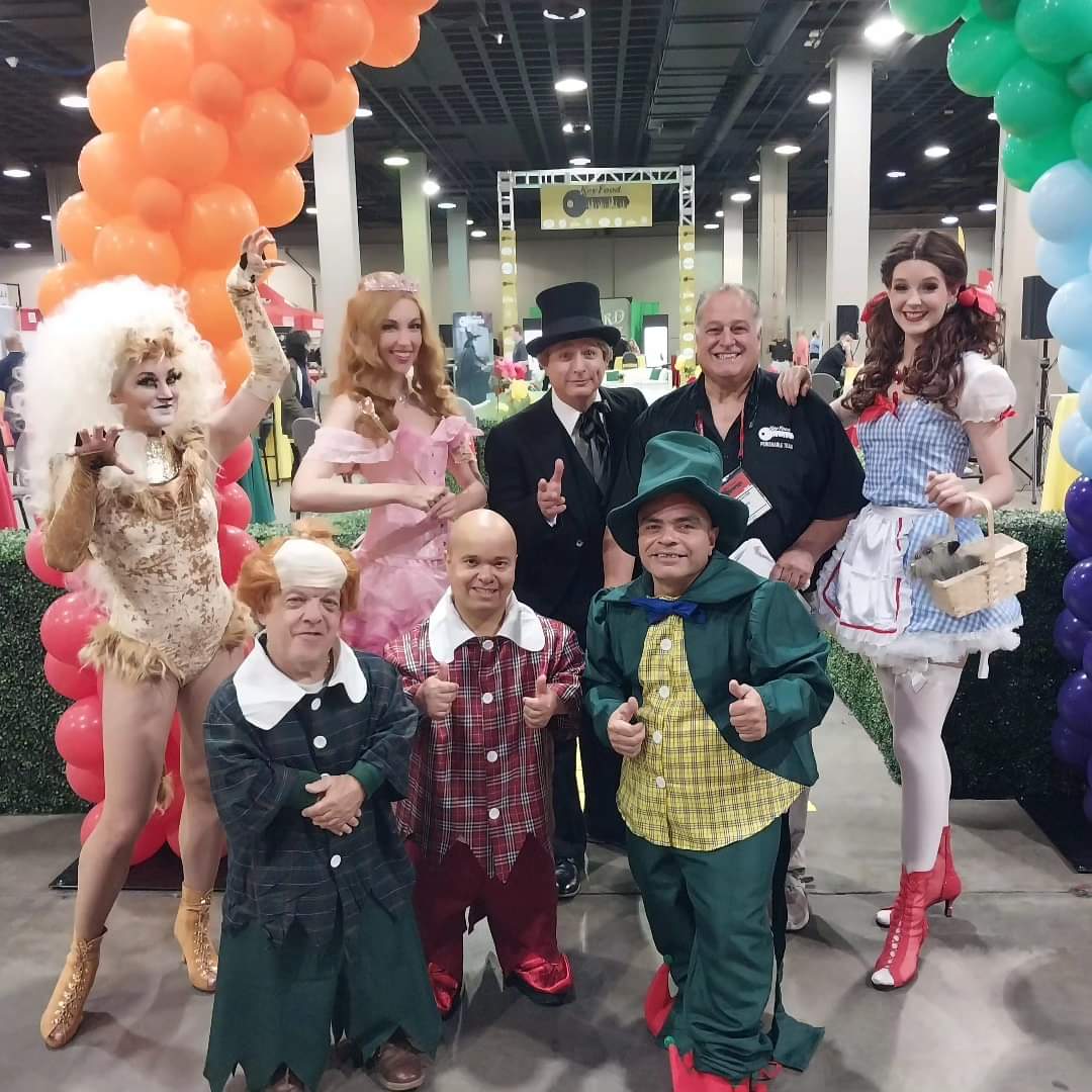 A group of eight people in colorful costumes poses indoors, surrounded by vibrant balloon arches.